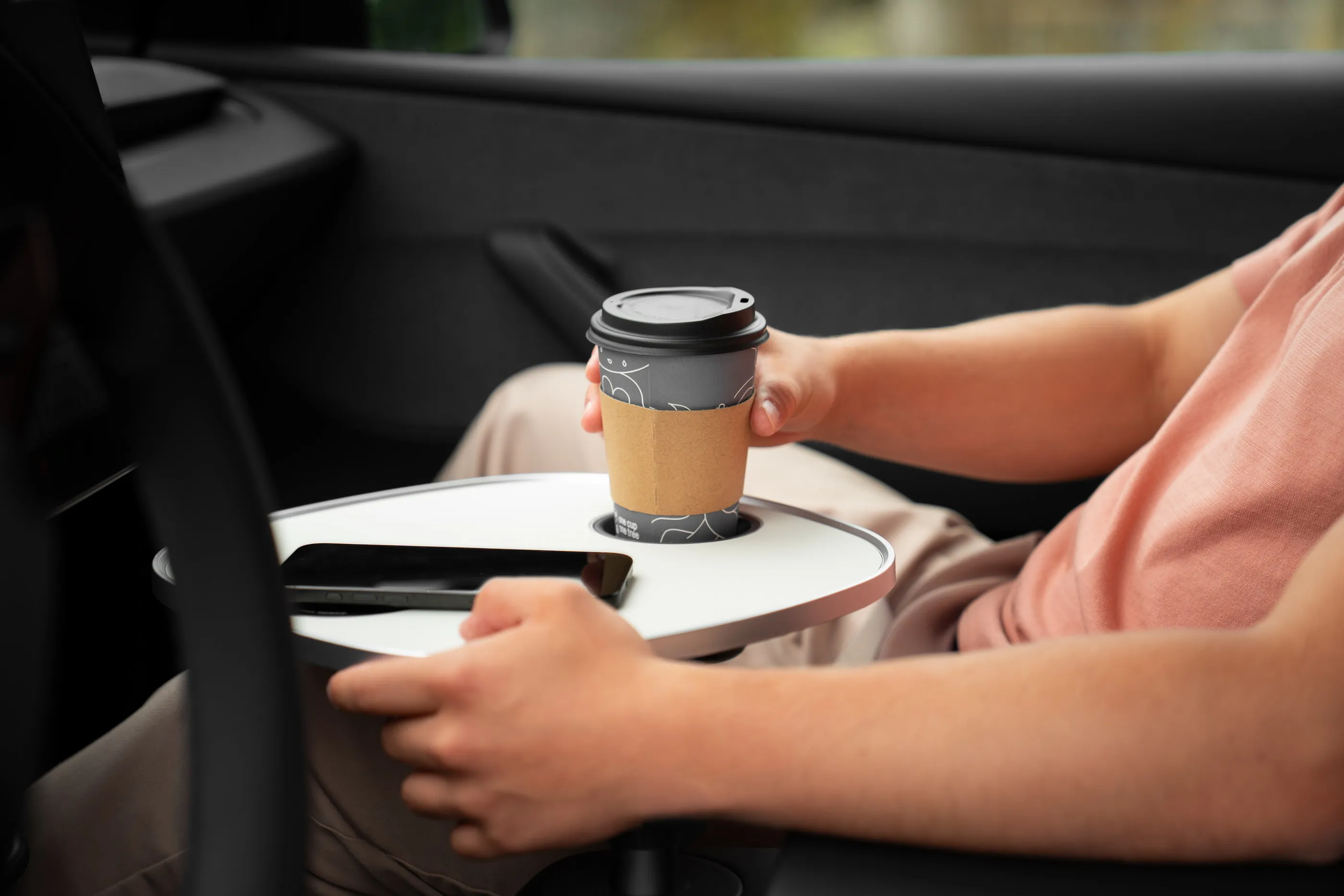 Person hält einen Kaffeebecher über der weißen Tischplatte, auf der ein Smartphone flach liegt