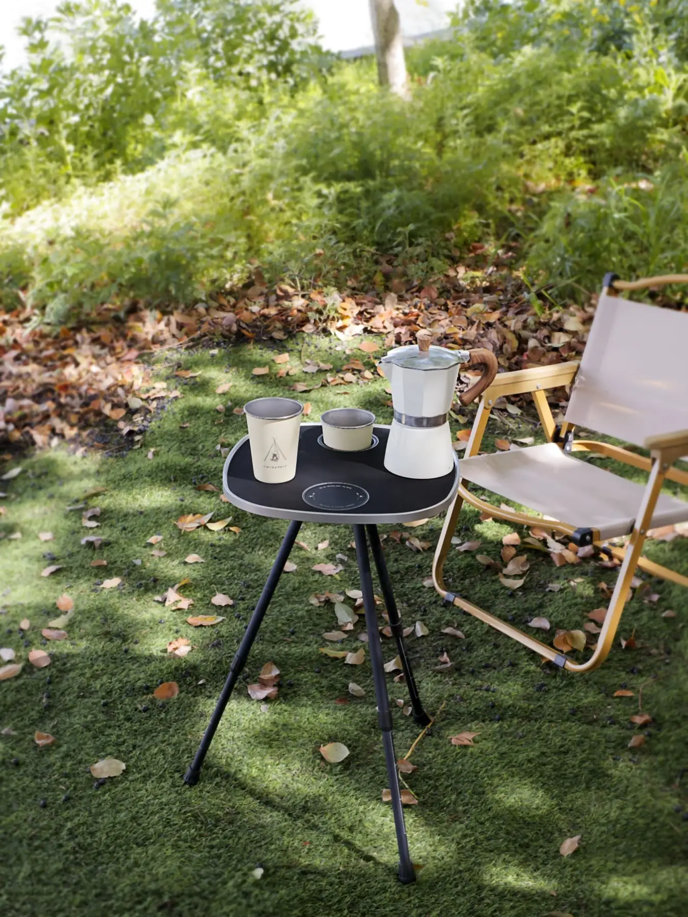 Schwarze Tischplatte auf den drei Teleskop-Standbeinen im Grünen, darauf eine Espressokanne und zwei Becher, daneben ein Campingstuhl